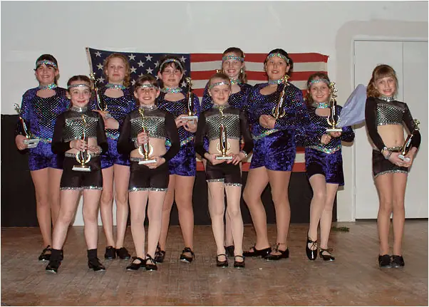 Ten children in sequined costumes pose indoors with trophies in front of an American flag.