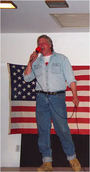 A man sings into a red microphone in front of a large American flag.