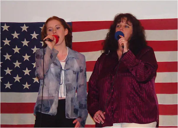 Two women sing into handheld microphones in front of a large American flag.