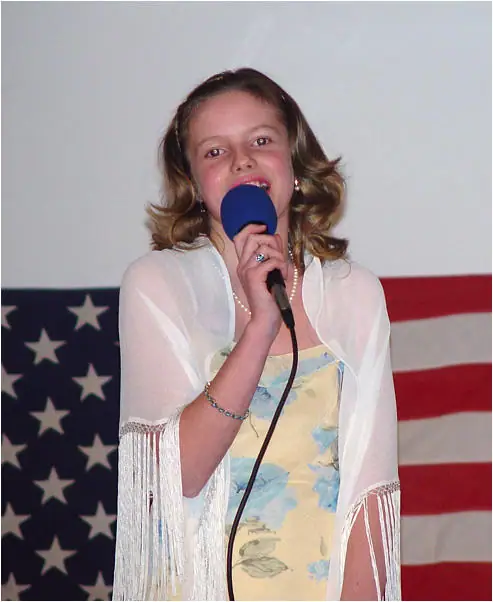 Young woman holding a microphone in front of a large American flag.