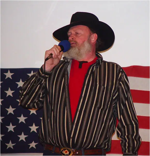 Bearded man in a cowboy hat speaks or sings into a microphone before an American flag.