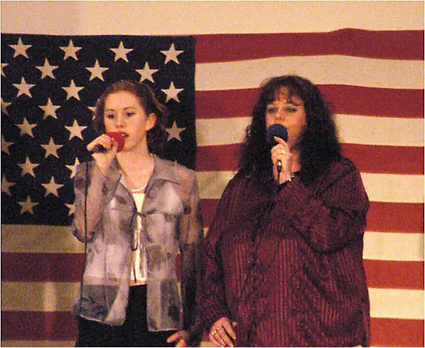 Two women sing into microphones in front of a large American flag.