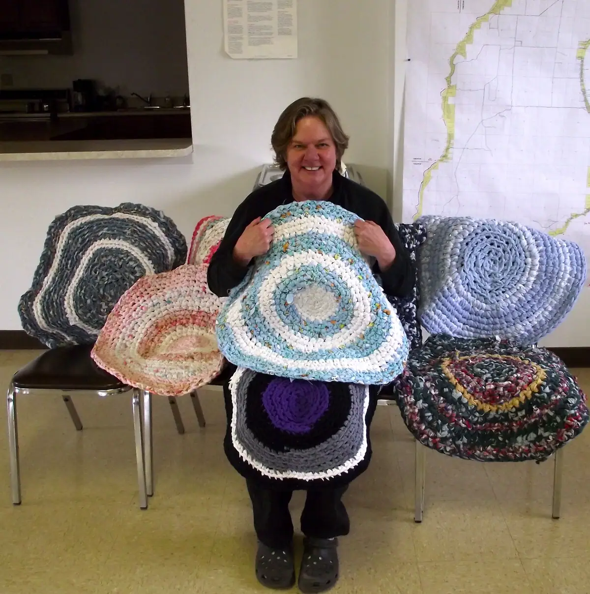 A smiling woman holds a blue-and-white circular rag rug among several handmade rugs on chairs.