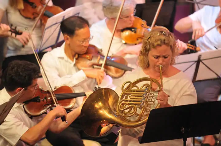 Musicians perform together, including violinists and a French horn player reading from sheet music.