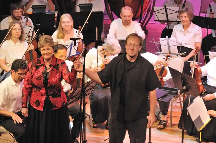 A man and woman stand before seated musicians holding string instruments and music stands.