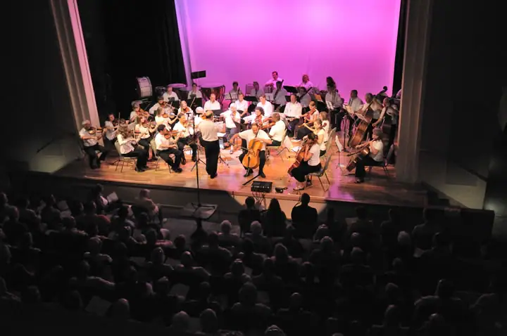 An orchestra performs on a brightly lit stage before a seated audience.