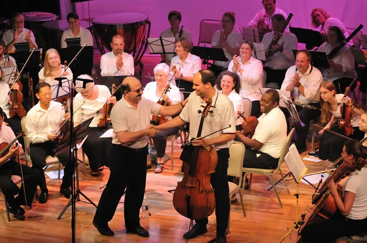 Musicians sit on a brightly lit stage as two men shake hands beside a cello.