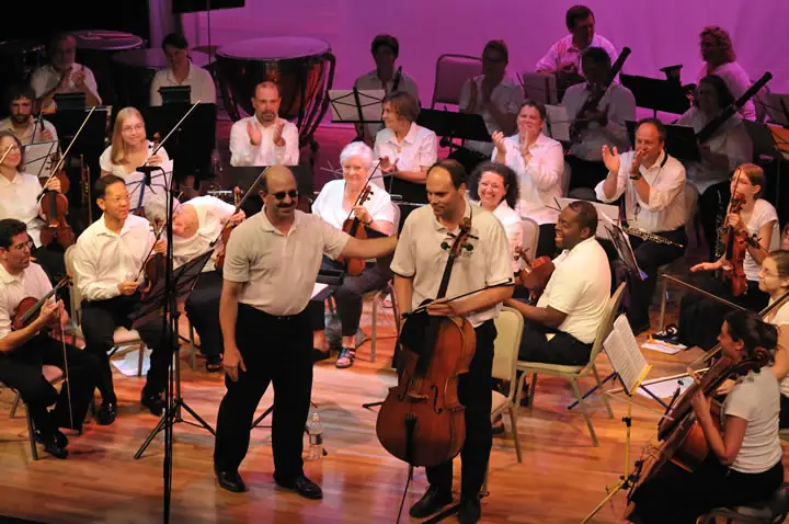 Musicians perform on a brightly lit stage, with a cellist and another standing performer in the foreground.