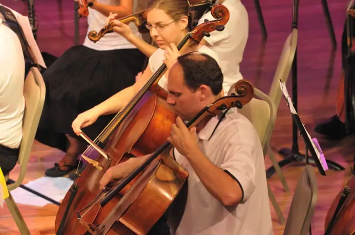 Seated musicians perform on cellos and other string instruments, with music stands and chairs around them.