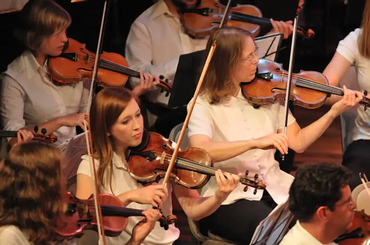 Several violinists in white shirts perform together, their bows moving across the strings.