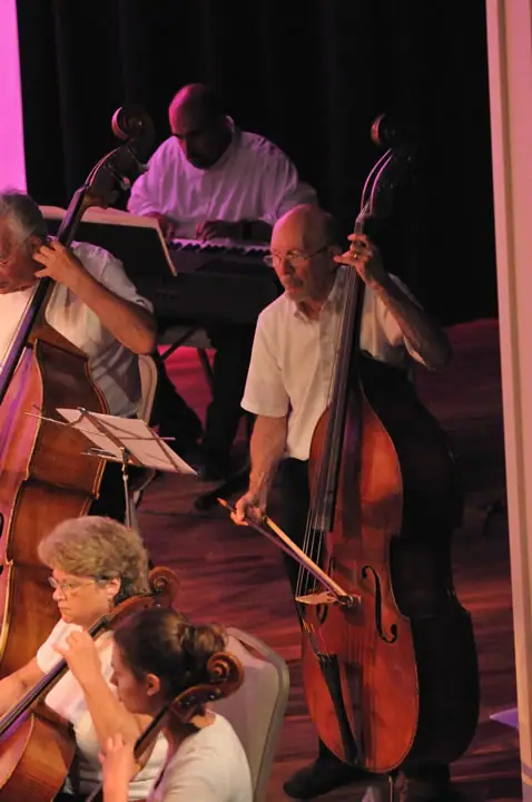 Musicians perform onstage with piano, double basses, cellos, and sheet music.