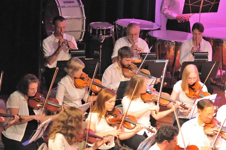 Musicians play violins and brass instruments on a brightly lit stage behind music stands.