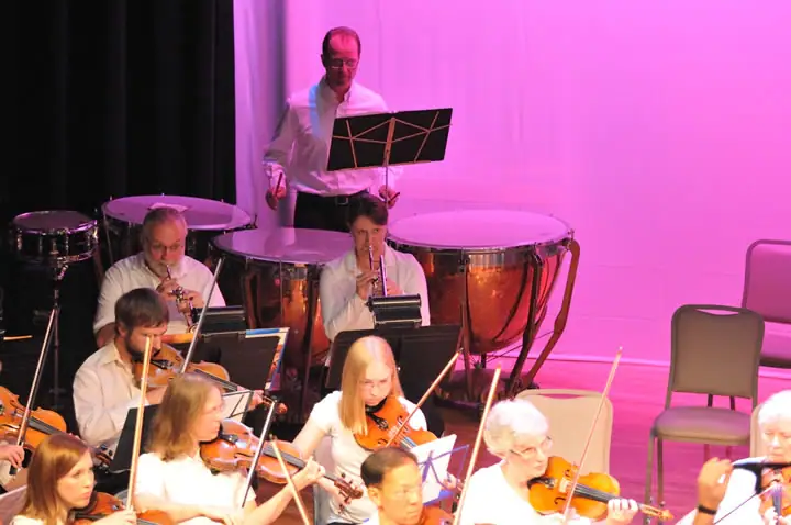 Musicians perform on a brightly lit stage with violins, music stands, and large timpani drums.