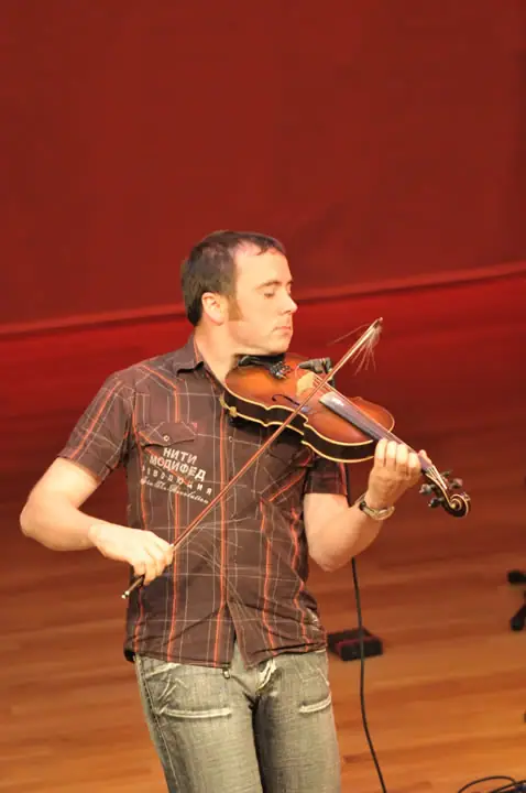 A man plays a violin and bow against a red wood-paneled stage backdrop.