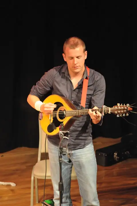 A seated musician plays a mandolin on a small stage beside a microphone.