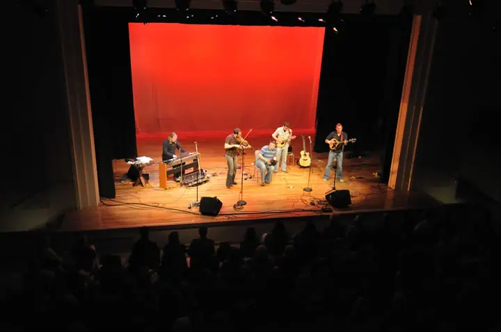 Five musicians perform on a theater stage before a seated audience.