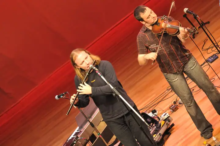 Two musicians perform with a flute and violin on a stage with microphones and a red curtain.