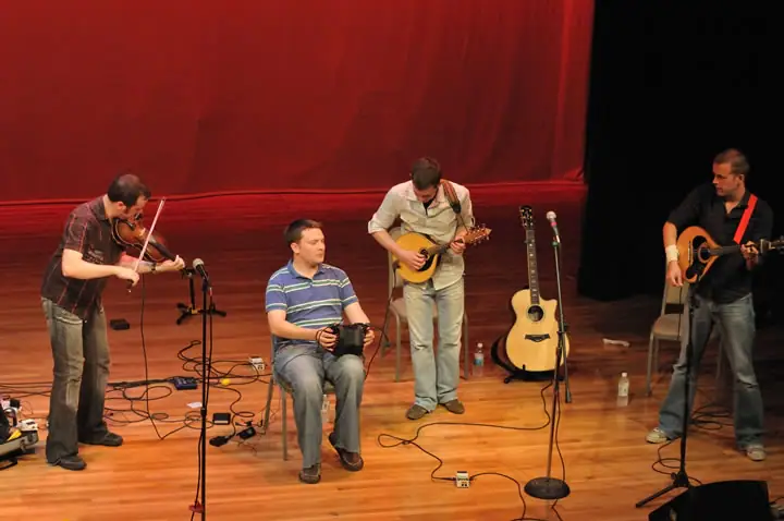 Four musicians perform on a wooden stage before a red curtain, with string instruments, microphones, and cables visible.