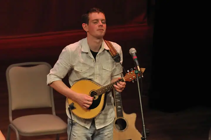 A musician plays a mandolin beside a microphone on a dimly lit stage.