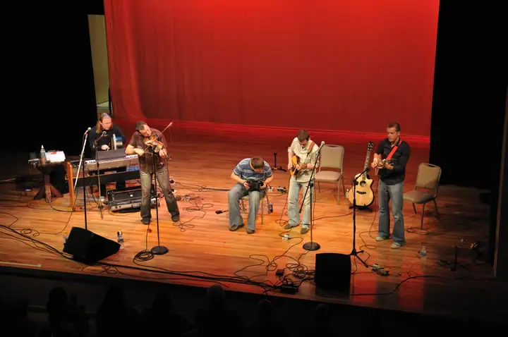 Five musicians perform on a theater stage before a seated audience, with instruments, microphones, and cables visible.