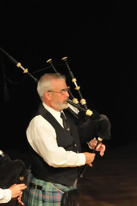 A bagpiper in a tartan kilt performs beside another partially visible musician.