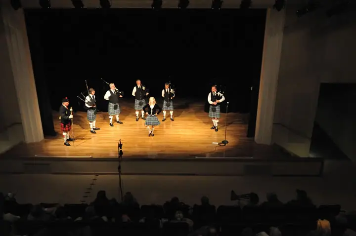 A pipe band performs on a theater stage before a seated audience.