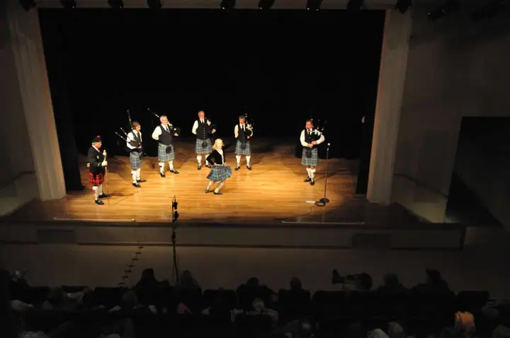 A pipe band and dancer perform on a lit theater stage before a seated audience.