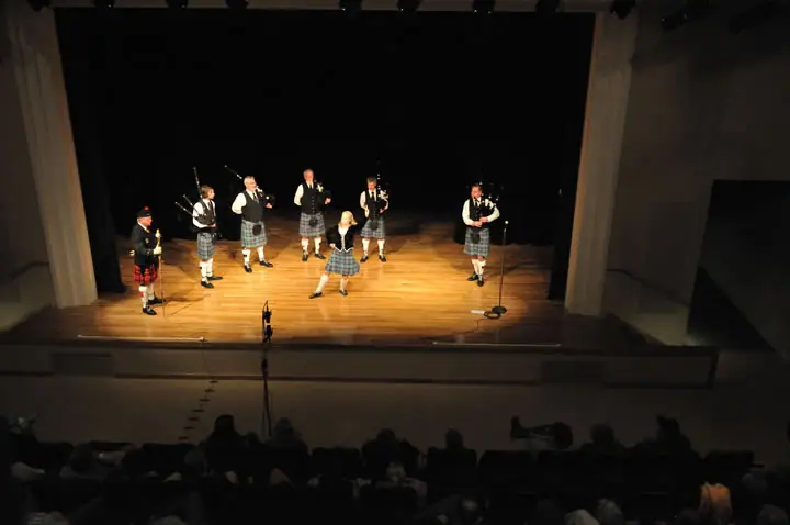 Bagpipers and a dancer perform on a lit stage before a seated audience.