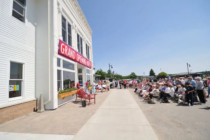 A crowd gathers beside a white building beneath a banner reading “GRAND OPENING JULY.”
