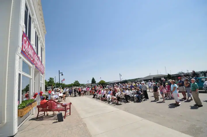 A large outdoor gathering takes place beside a building displaying a GRAND OPENING banner.
