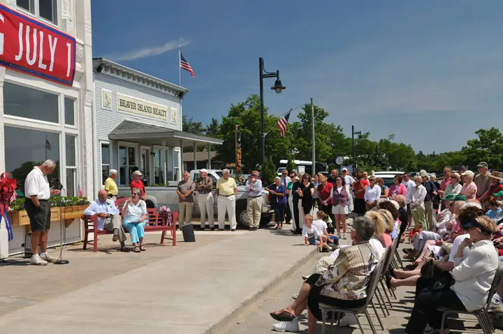 Outdoor ceremony with a speaker, seated audience, flags, and a building marked “BEAVER ISLAND REALTY.”