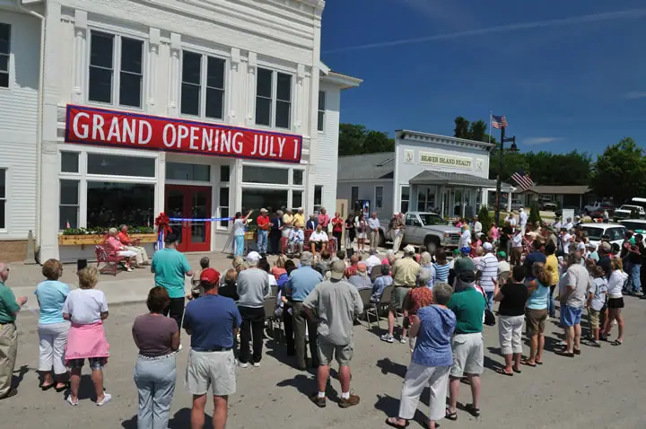 A crowd gathers outside a white building beneath a red “GRAND OPENING JULY 1” banner.