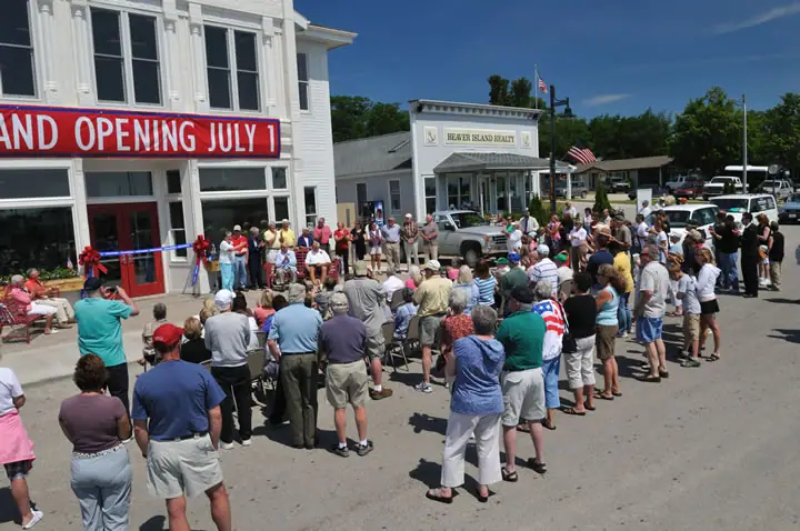 A large crowd gathers outside a storefront for a ribbon-cutting ceremony beneath a grand-opening banner.