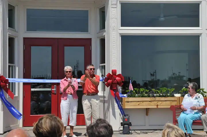 Two adults cut a blue ribbon before a building entrance as spectators watch.