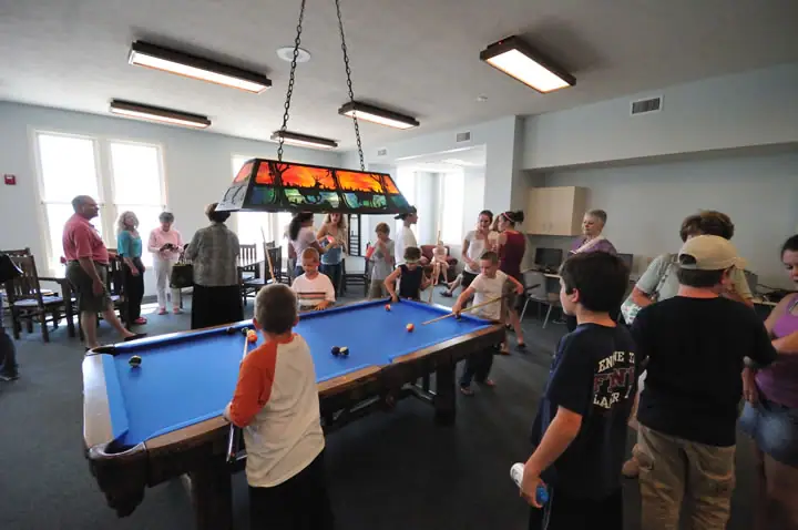 Adults and children gather around a blue-felt pool table beneath a stained-glass light fixture.