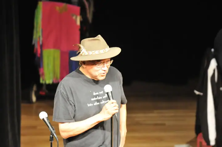 A man in a feathered hat holds a microphone on a wooden stage.