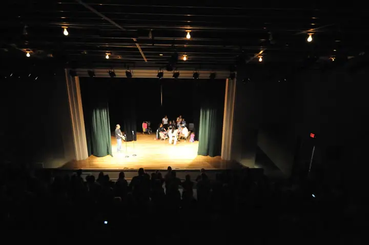 A brightly lit stage with seated participants and a standing speaker viewed from a dark auditorium.