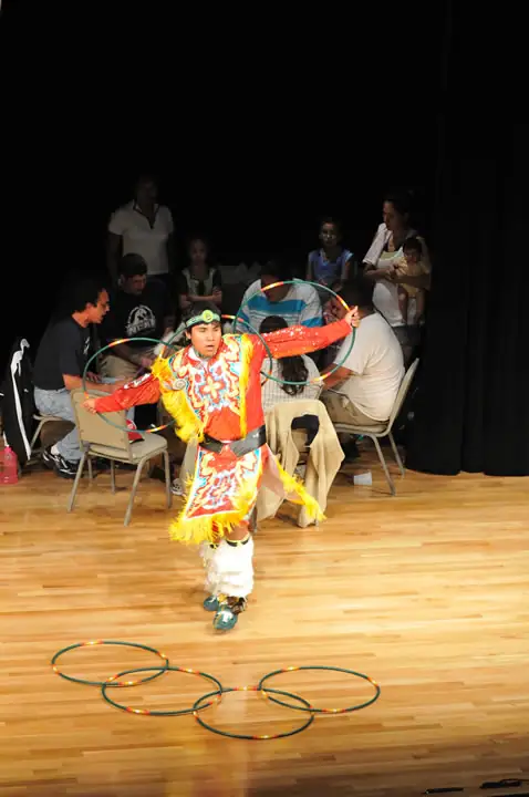 Performer in a colorful fringed outfit holds hoops on a wooden stage before a seated audience.
