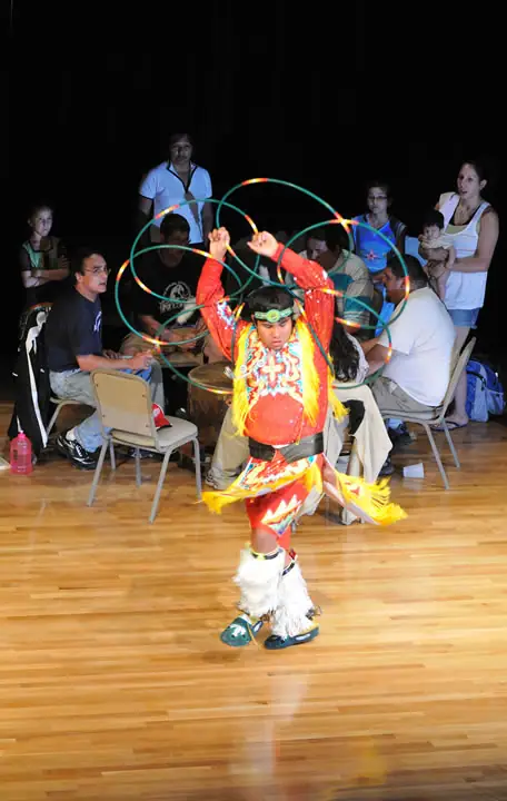 A dancer in colorful regalia performs with several hoops before a seated audience.