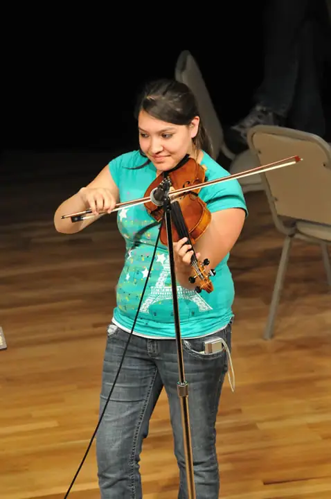 Young violinist performing indoors with a bow, microphone stand, and folding chairs behind her.
