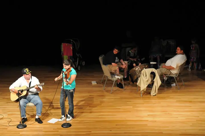 Two musicians perform on a wooden stage while seated people watch from folding chairs.