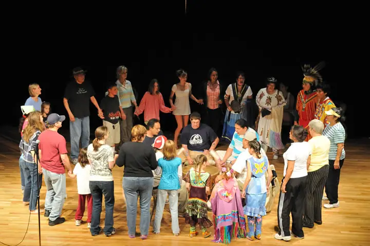 Adults and children gather in a circle on a wooden stage, with several wearing colorful regalia and feathered headdresses.