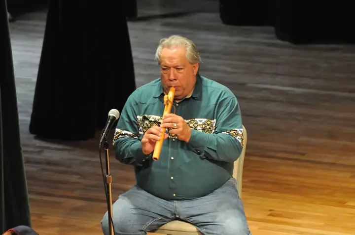 Seated musician playing a wooden flute on a stage beside a microphone.