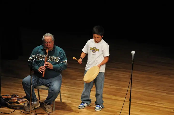 Two musicians perform on a wooden stage, with an adult playing flute and a child holding a frame drum.