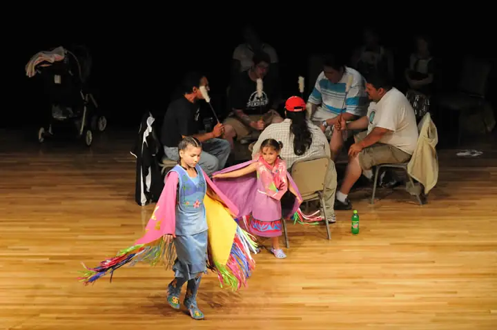 Two children in colorful fringed outfits walk across a wooden gym floor as adults sit behind them.