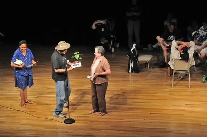 Adults gather on a wooden stage, with one person speaking into a microphone while others hold papers and a plant.
