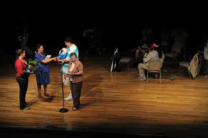 Adults gather around a microphone on a stage while others sit in the background.