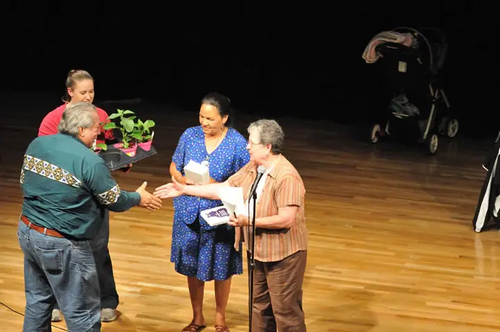 Four people gather on a stage as a man shakes hands with a woman holding papers and another person carries potted plants.