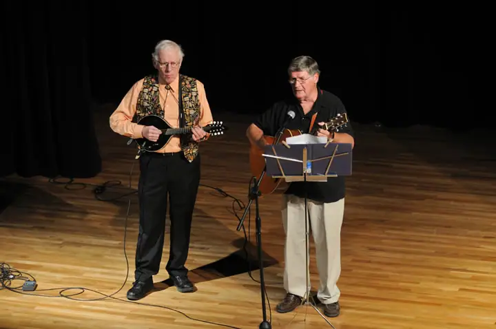 Two musicians perform on a wooden stage, playing a mandolin and an acoustic guitar beside microphones and a music stand.