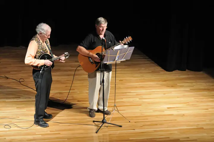 Two musicians perform on a wooden stage with a mandolin, acoustic guitar, microphone, and music stand.
